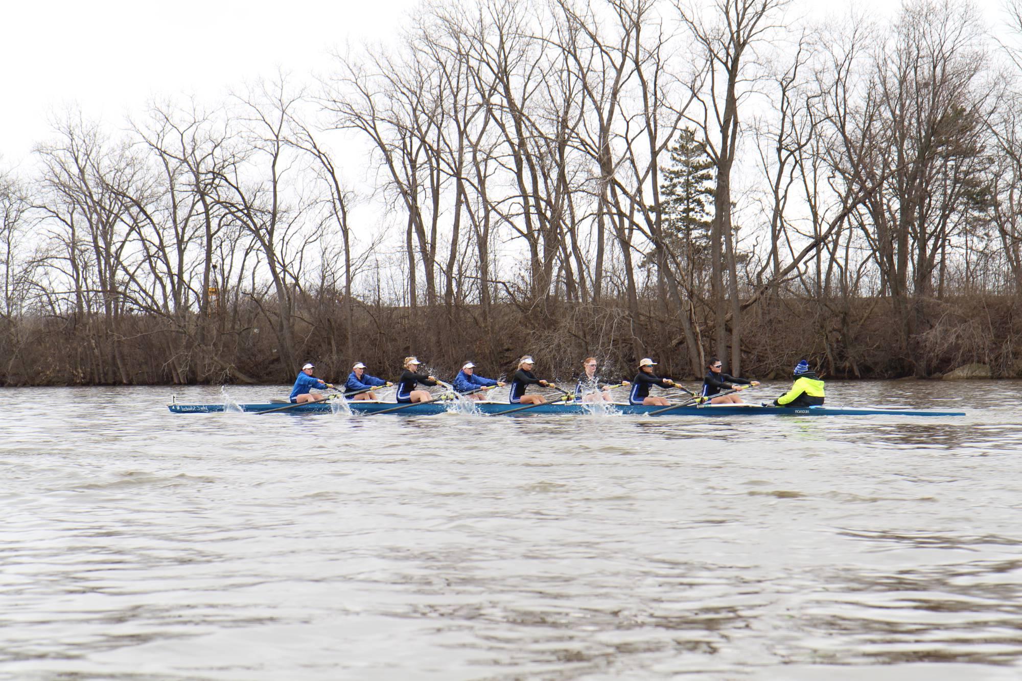 8 womens rowing during an overcast day on a river with trees with no leaves in the background